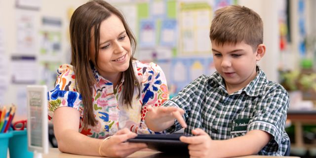 Teacher supporting a student using a tablet in the classroom, highlighting personalised learning, digital engagement and supportive guidance at St Ambrose’s Primary School